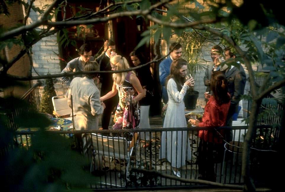 A group of people socializes on a patio surrounded by plants and trees. Some are holding drinks and chatting, while others gather around a table. The scene is lively and informal, viewed through leafy branches.