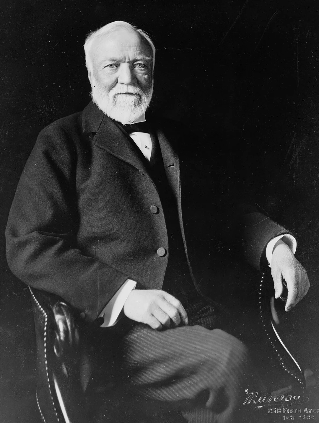 An older man with a white beard and mustache sits in a dark suit and vest, resting one hand on his knee and the other on the arm of a chair, looking slightly to the side against a dark background.