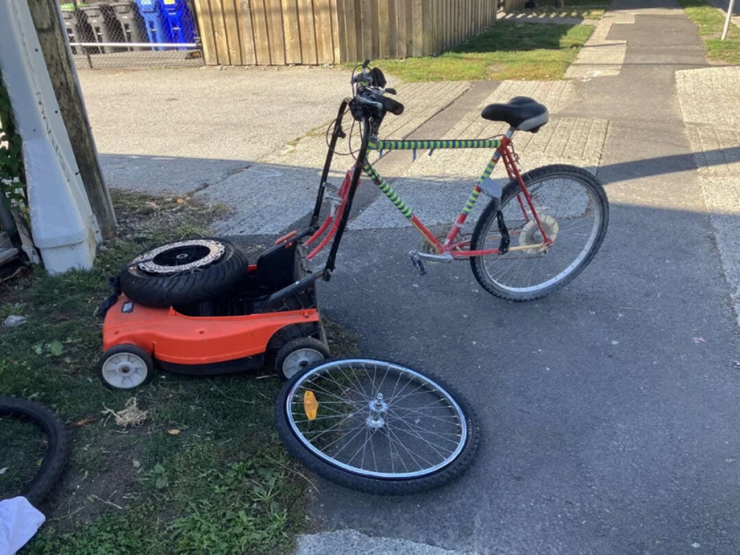 A red bicycle with a missing front wheel is attached to an orange lawn mower. The detached front wheel and another tire are lying on the ground nearby in a paved outdoor area.