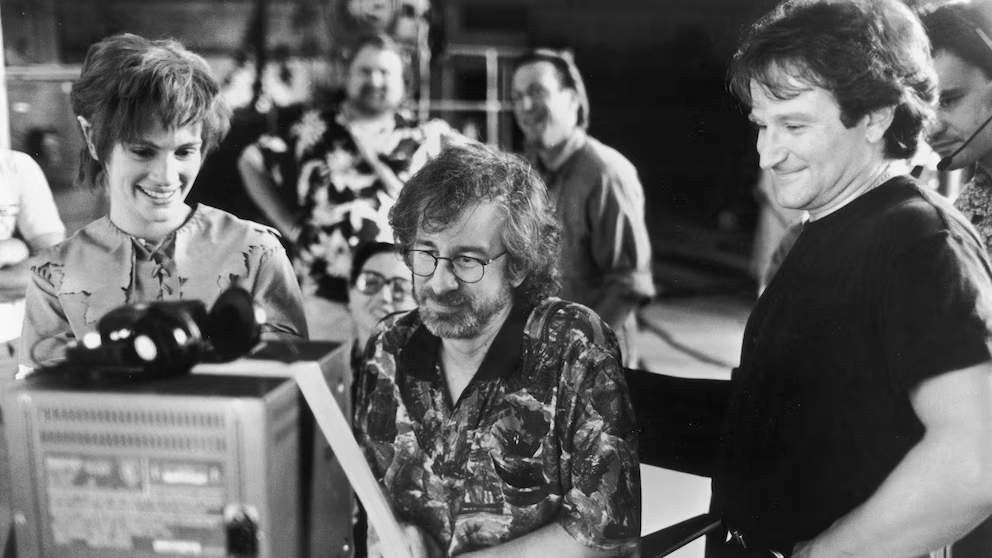 Three people look at a monitor on a film set, smiling and engaged. The setting appears informal and collaborative, with others in the background and filmmaking equipment visible around them.