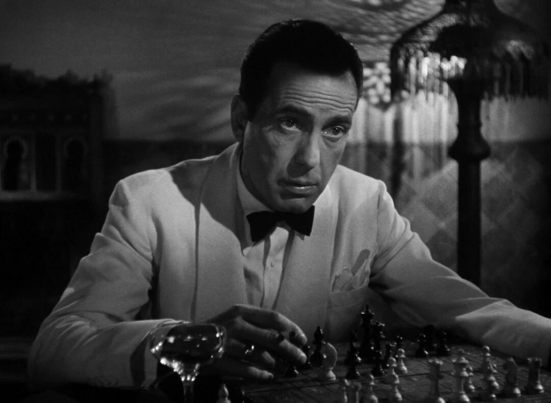 A man in a white tuxedo and black bow tie sits at a table playing chess, looking thoughtfully ahead. A glass and ornate lamp are visible in the dimly lit, vintage-style room.