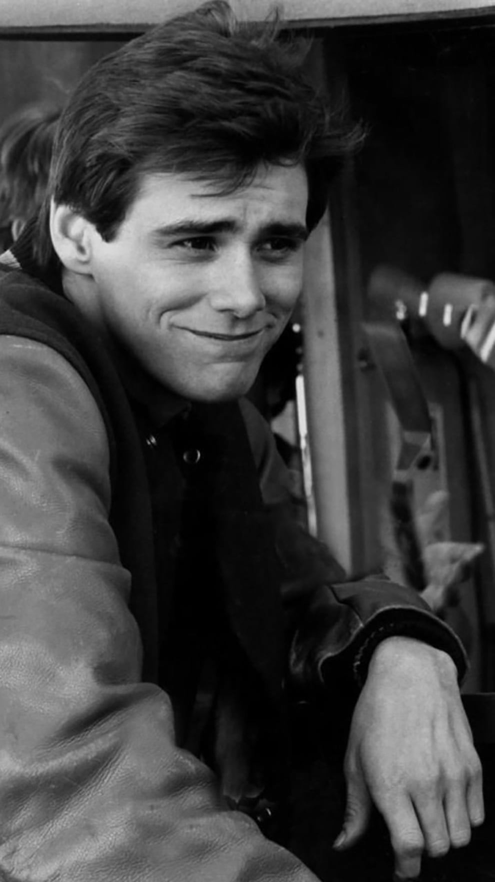 A young man with dark hair smiles softly while leaning on his arm, wearing a bomber-style jacket in a black and white photo.