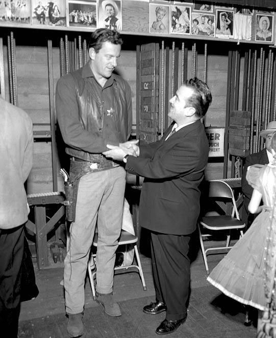 A very tall man in a cowboy costume shakes hands and smiles with a shorter man in a suit indoors, while others stand nearby. Photos are displayed on the wall behind them.