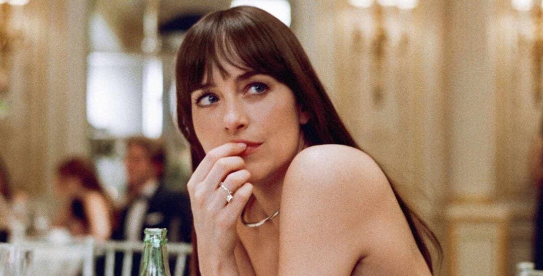 A woman with long brown hair and bangs sits at an elegant table, looking thoughtfully to the side with her hand near her mouth. A bottle and glass are in front of her, and the background is softly lit and blurred.