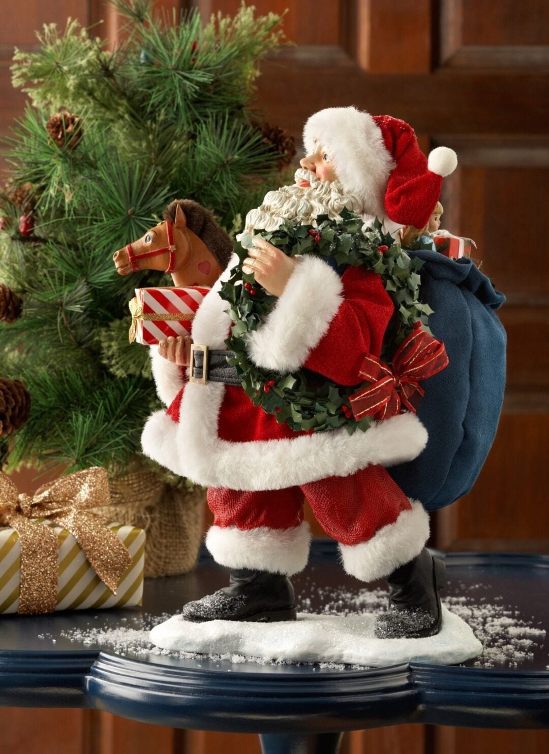 A festive Santa Claus figurine holding a wreath and toy horse, carrying a blue sack of gifts, stands on a snowy surface beside a decorated gift and pine tree branches with pinecones.