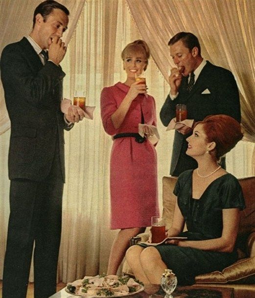 Four well-dressed adults enjoy drinks and hors d'oeuvres at an elegant indoor gathering; two men in suits stand with drinks and napkins, while two women in dresses, one seated, smile and converse.