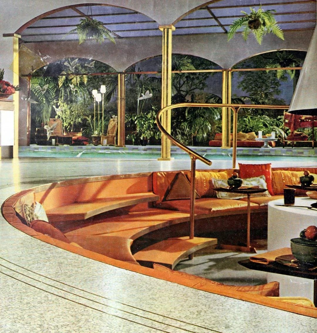 A retro sunken living room with orange cushioned seating surrounds a white circular table. The area overlooks a pool and lush greenery, with gold railings, arched windows, and hanging plants adding a luxurious, vintage feel.