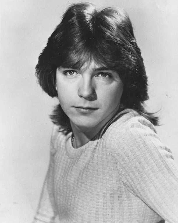 Black-and-white portrait of a young man with medium-length, feathered hair, wearing a light-colored, textured sweater, and looking directly at the camera with a neutral expression.