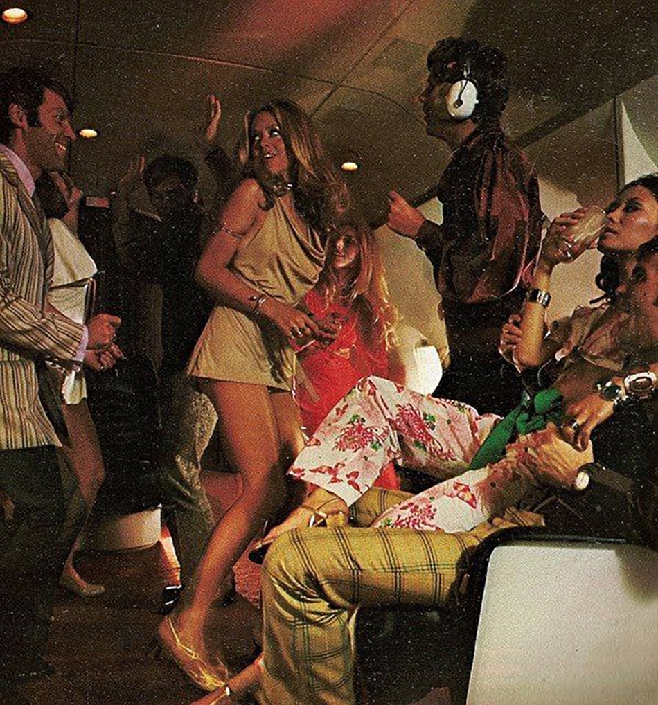 A group of men and women in 1970s-style clothing dance and socialize inside an airplane cabin, with some people seated and others standing, drinks in hand and lively expressions.