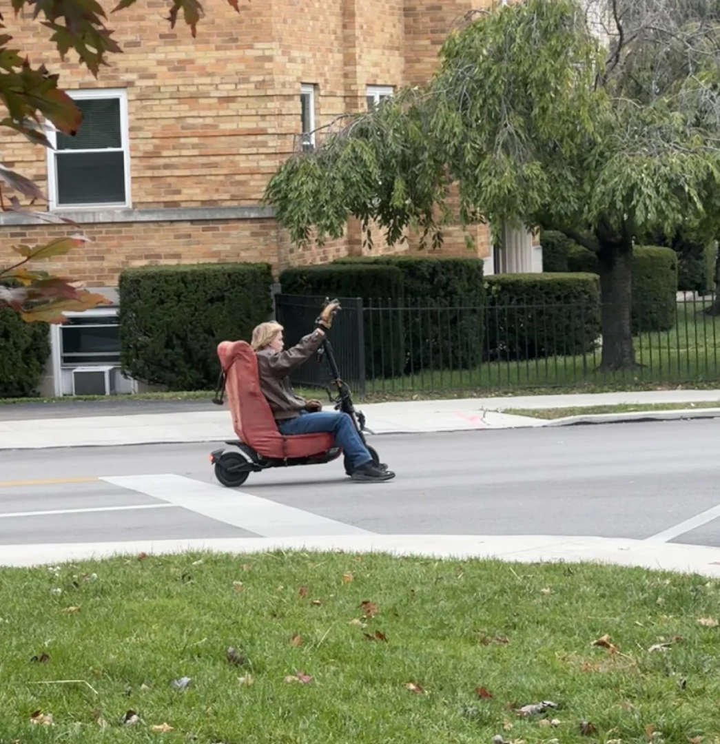 A person rides a motorized scooter with a red upholstered armchair as a seat, traveling down a suburban street lined with grass, trees, and brick buildings.