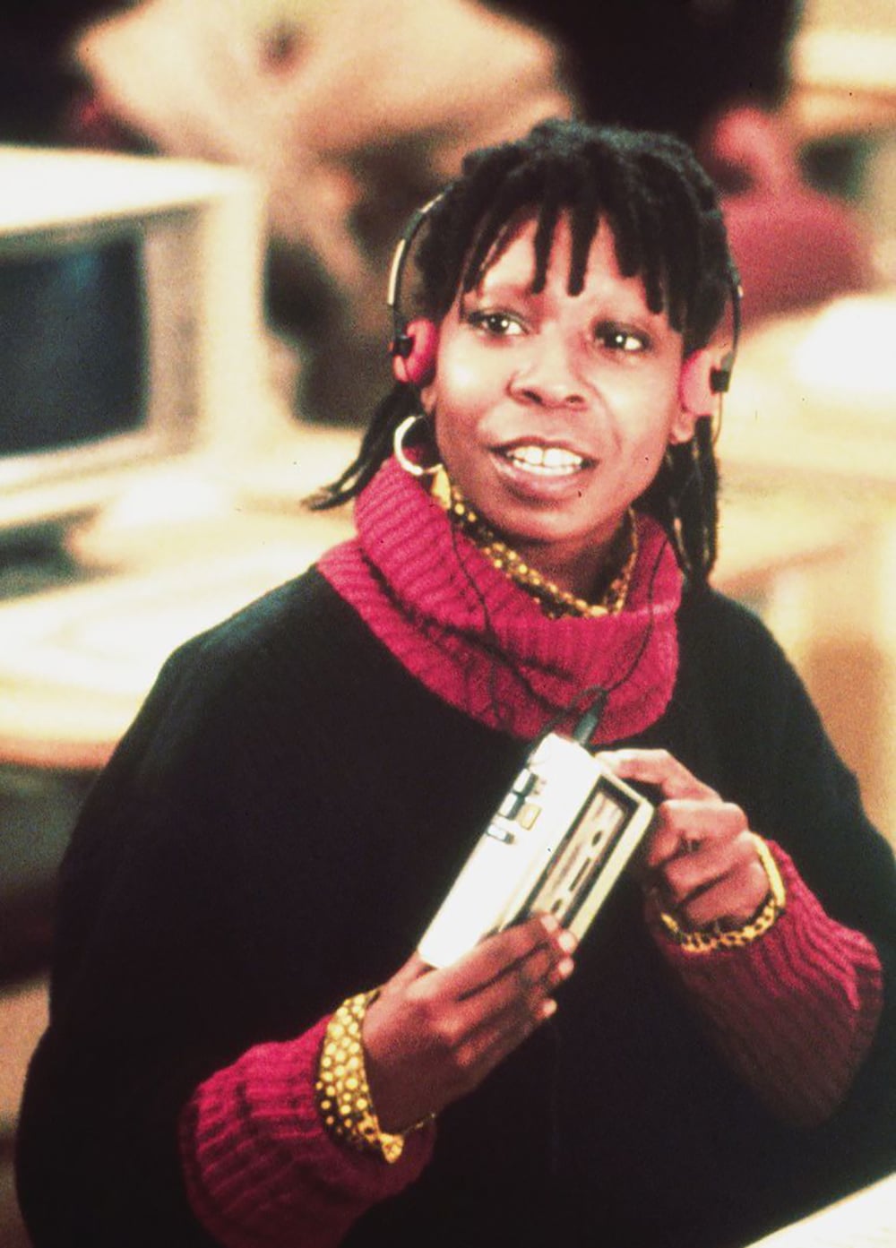 A person wearing a red turtleneck sweater, black vest, and headphones holds a portable cassette player. They are smiling, and an old computer monitor is visible in the background.