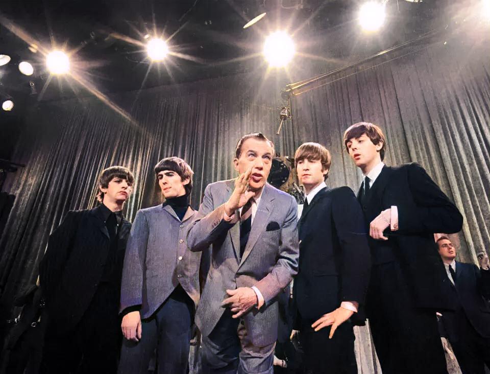 A man in a suit stands in front of four young men wearing suits on a stage with bright lights and a curtain backdrop. The man gestures while the others stand close, looking in different directions.