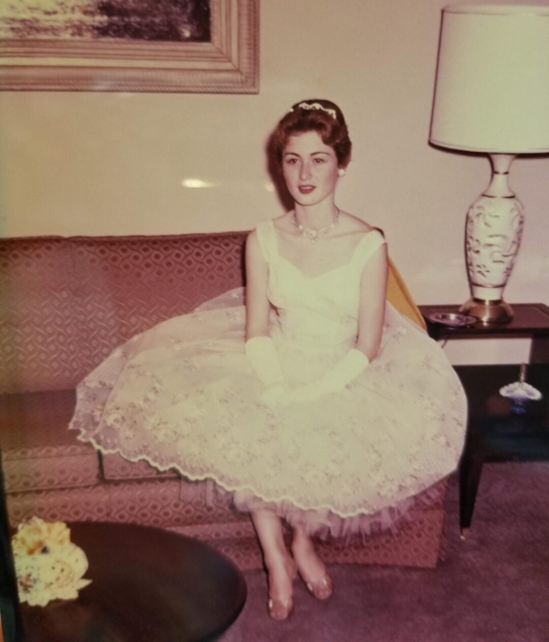 A young woman in a formal white dress, gloves, and ballet flats sits on a patterned couch beside a table lamp in a living room, with a framed painting on the wall behind her.