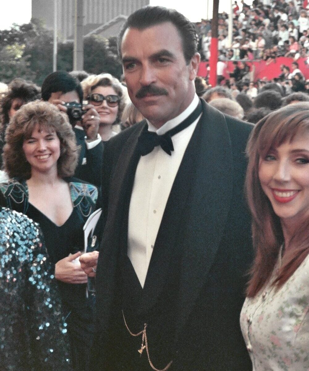 A man in a black tuxedo stands smiling at an outdoor event with a crowd and photographers in the background. He has a mustache and is accompanied by two women, one with curly hair and one with straight hair, both smiling.