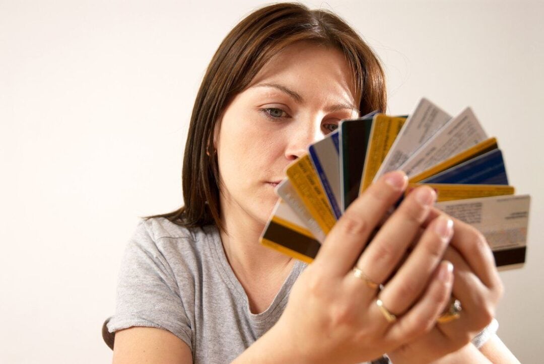 A woman wearing a gray shirt holds and examines a fanned-out stack of credit cards in her hands, looking at them thoughtfully against a plain background.