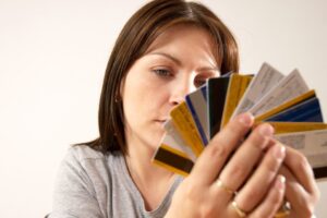 A woman in a gray shirt holds and examines a fan of various credit cards, looking at them thoughtfully. The background is plain and light-colored.