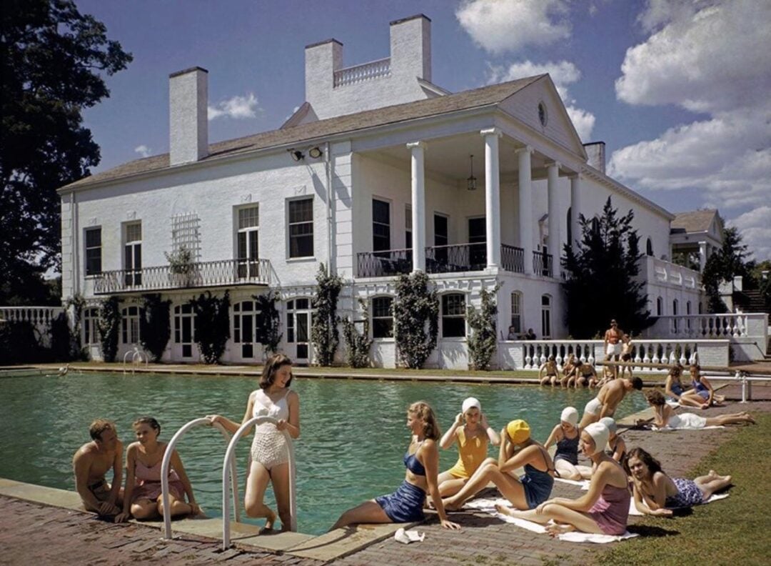 People in vintage swimsuits relax and socialize by a large pool in front of a grand white mansion with columns on a sunny day; some are sitting poolside, others stand in the water.