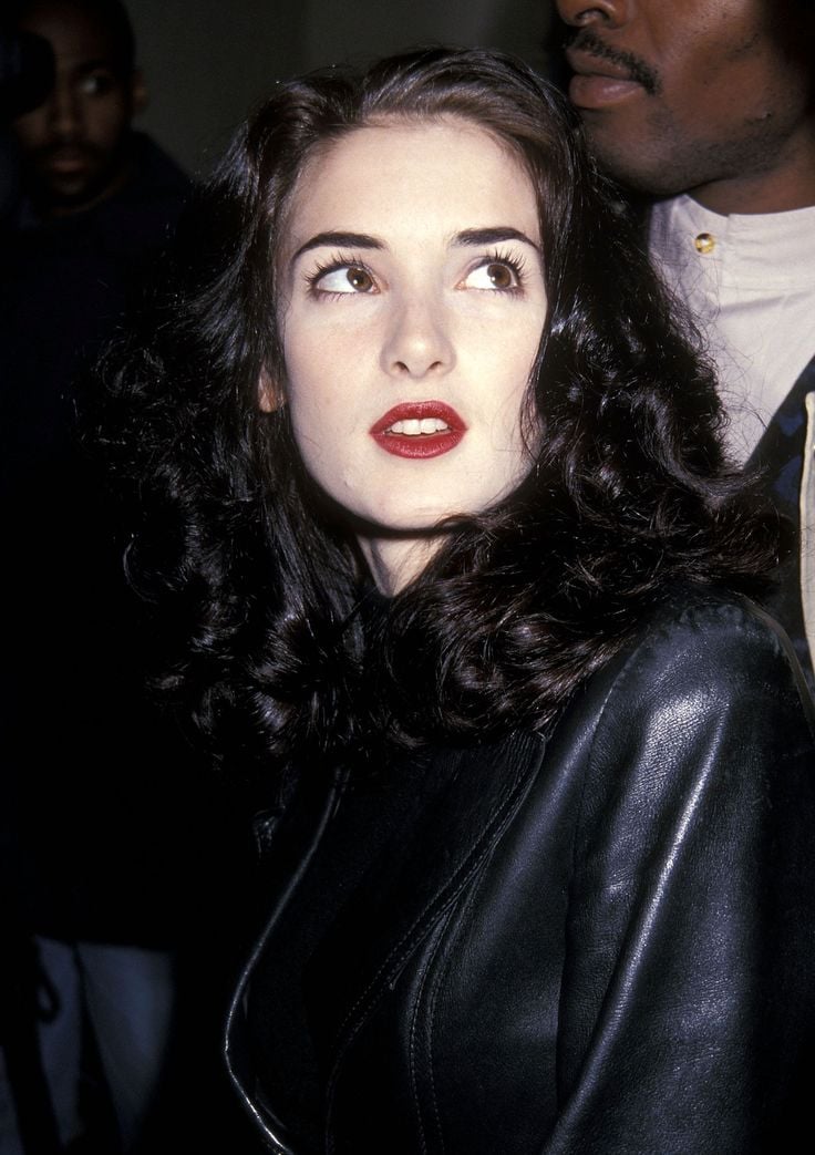 A woman with pale skin, wavy dark hair, and red lipstick wears a black leather jacket. She looks slightly upward and to the side, surrounded by people in a dimly lit environment.