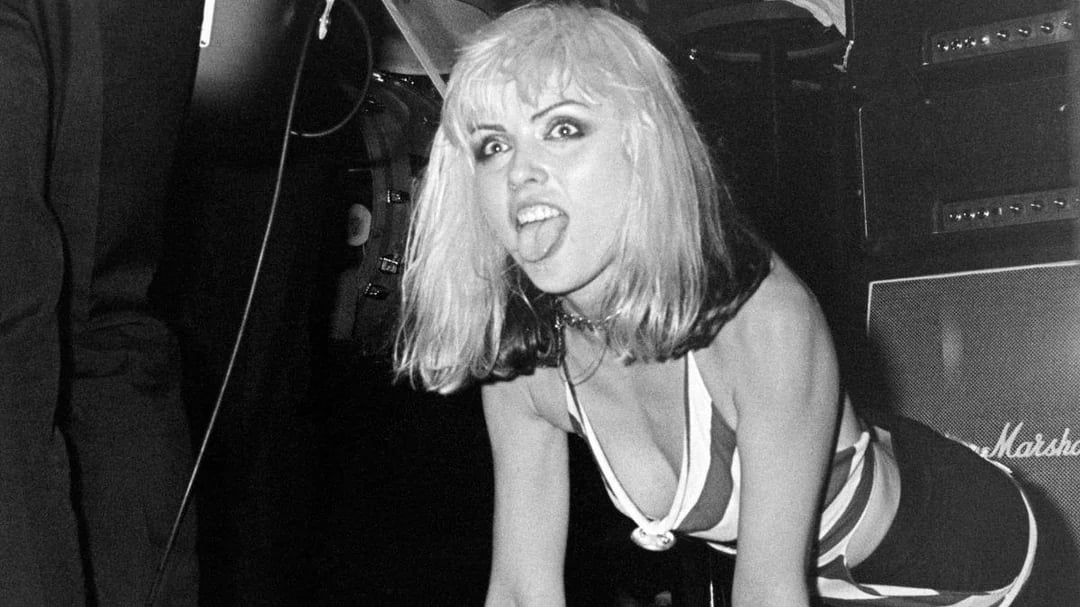 A woman with light hair and dark roots sticks out her tongue while crouching onstage, wearing a striped sleeveless outfit. Musical equipment is visible in the background. The photo is black and white.