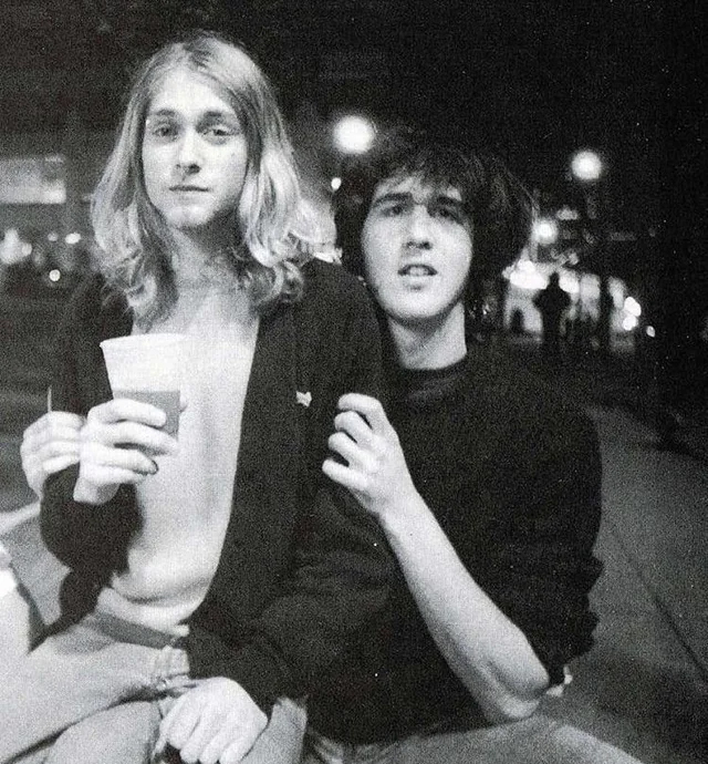 Two young people sit close together at night; one with long blond hair holds a cup and looks at the camera, while the other rests a hand on their shoulder. The background is an outdoor urban setting with blurred lights.