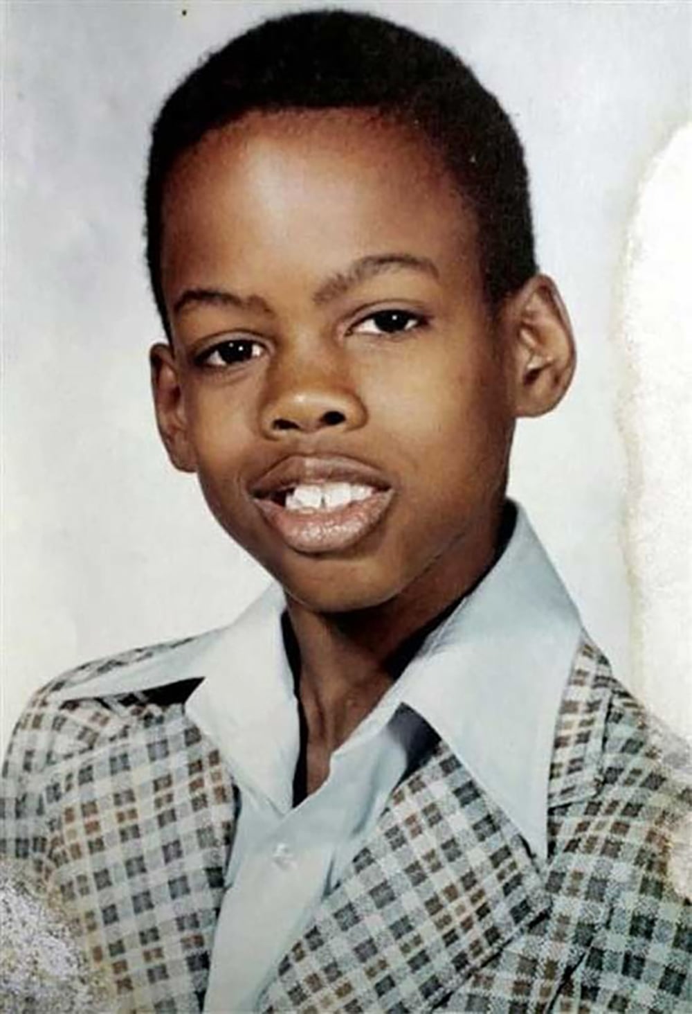 A young boy smiles at the camera, wearing a plaid jacket and a light blue collared shirt. He has short hair and is posed in front of a plain background, likely for a school portrait.