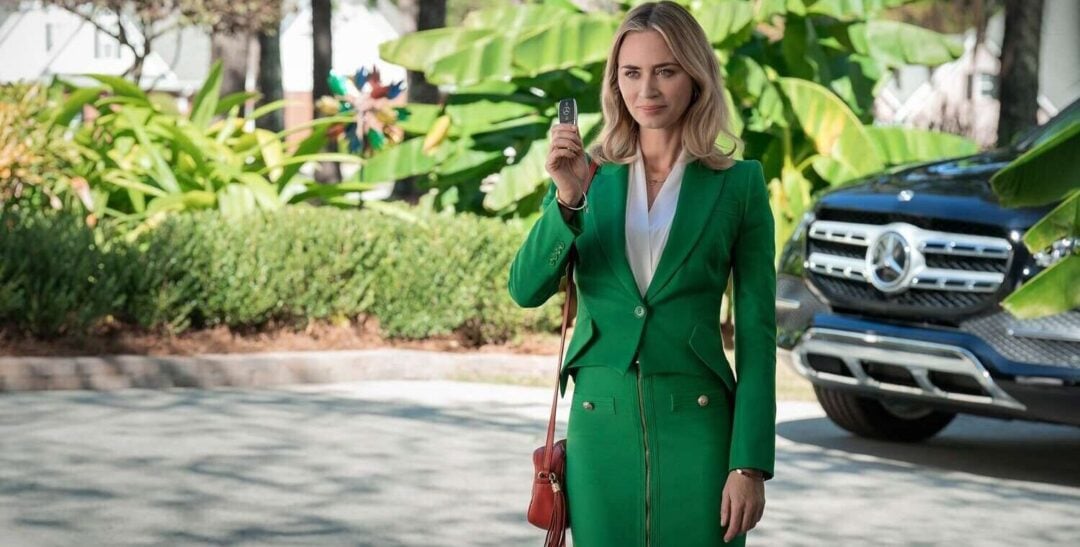 A woman in a bright green suit stands outdoors, holding up a car key. She has long blonde hair and carries a red purse. Behind her is a black Mercedes-Benz SUV and lush green plants.