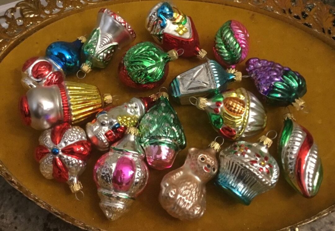 A gold tray holds assorted vintage-style glass Christmas ornaments in various shapes, colors, and designs, including a teddy bear, Santa, and bell. The ornaments are shiny and festive.