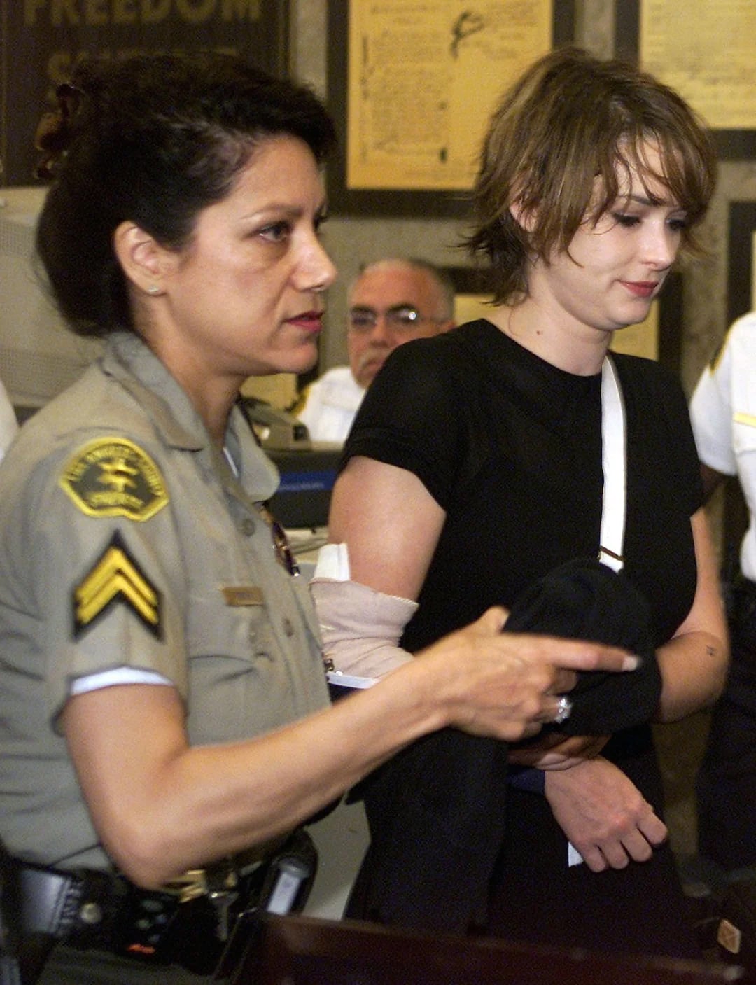 A uniformed officer escorts a woman with a short haircut and a bandaged arm through a room. The woman looks down, holding dark clothing, while people and framed documents are visible in the background.