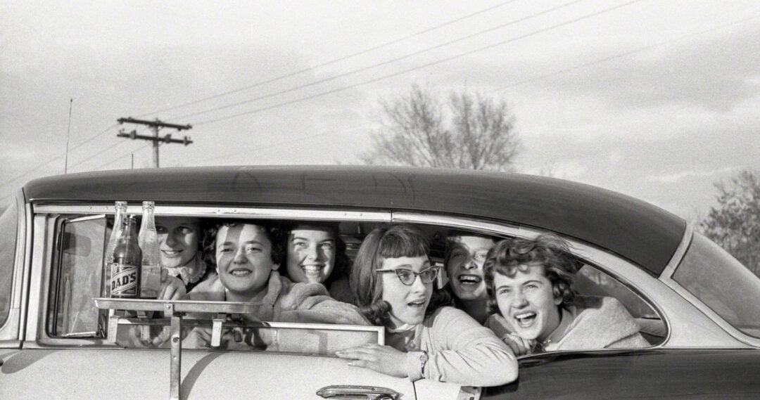 A group of six smiling young women lean out of the open window of a classic car. Two glass soda bottles rest on the car door, and the background shows trees and utility poles under a cloudy sky.