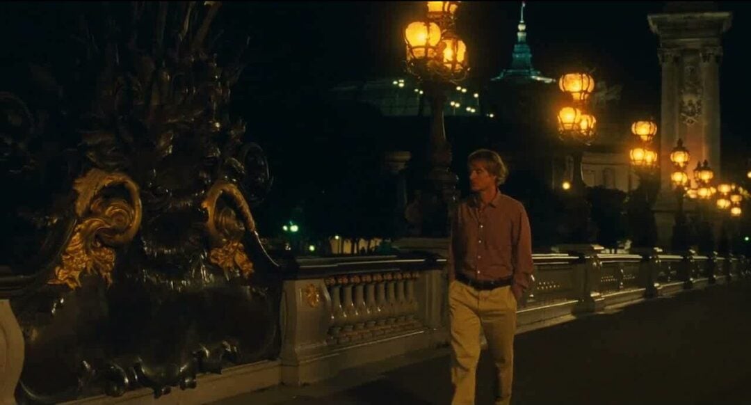 A man in a brown shirt and beige pants walks alone at night on a beautifully lit bridge adorned with ornate lanterns and gold details. City lights and a domed building are visible in the background.