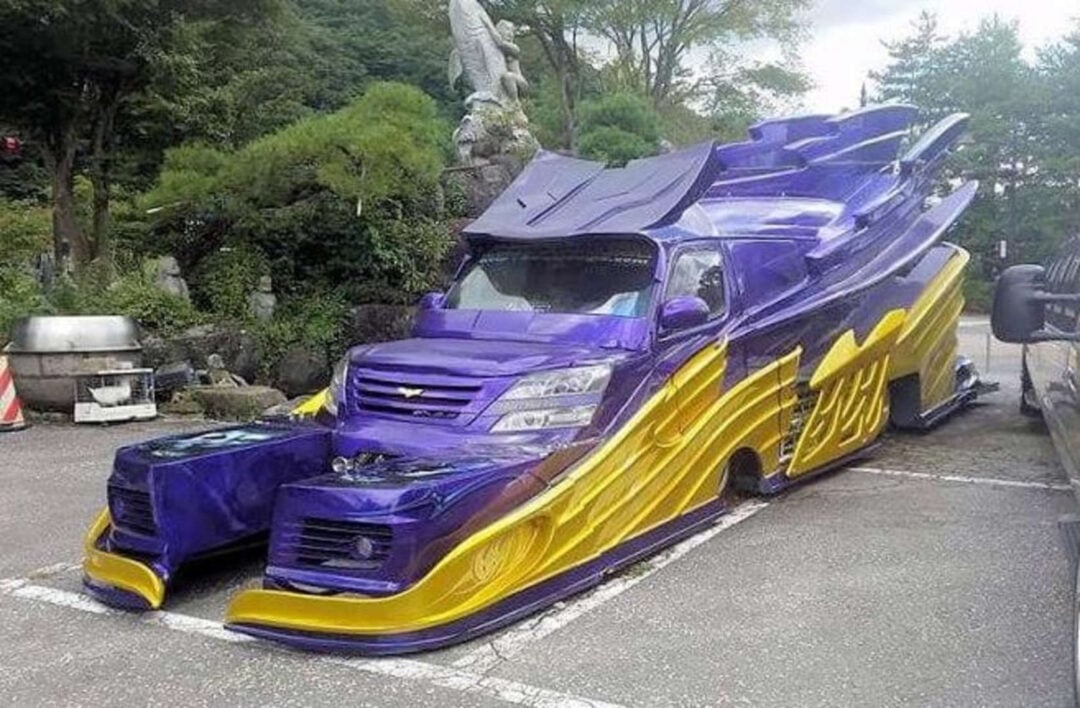 A highly customized car with exaggerated, futuristic bodywork in purple and yellow, featuring large fins, extended fenders, and a dramatic, angular design, parked in an outdoor lot surrounded by greenery.