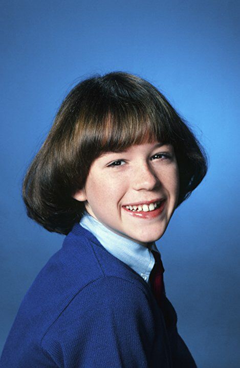 Smiling child with short brown hair and bangs, wearing a blue sweater over a collared shirt, against a plain blue background.