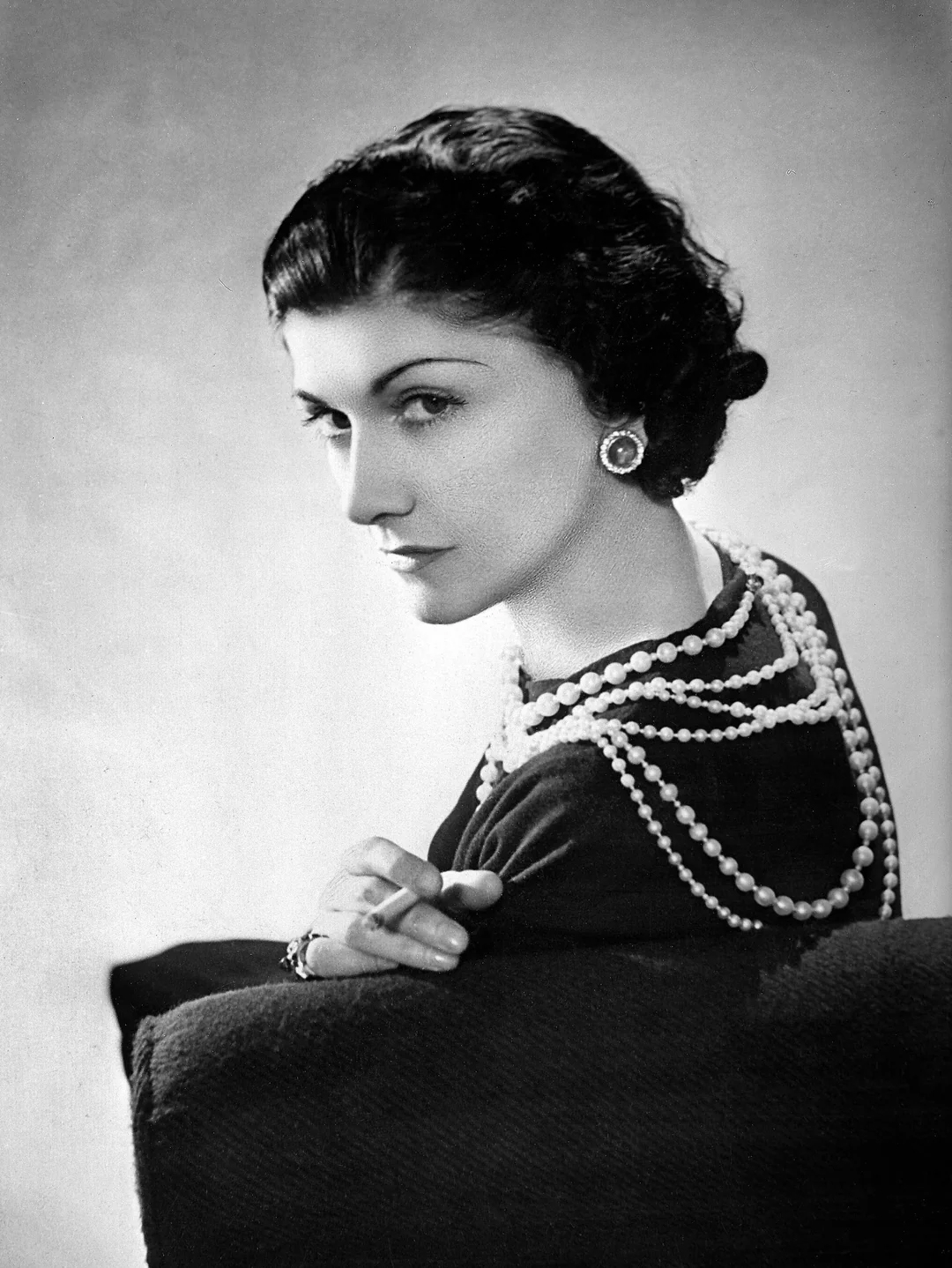 Black and white portrait of a woman with short dark hair, wearing pearl necklaces and earrings, seated sideways, glancing at the camera with a composed expression.