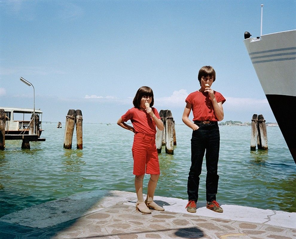 Two children wearing red clothes stand by the water, eating ice cream. The sky is clear and blue, and a large boat is partially visible on the right. Wooden posts emerge from the water in the background.