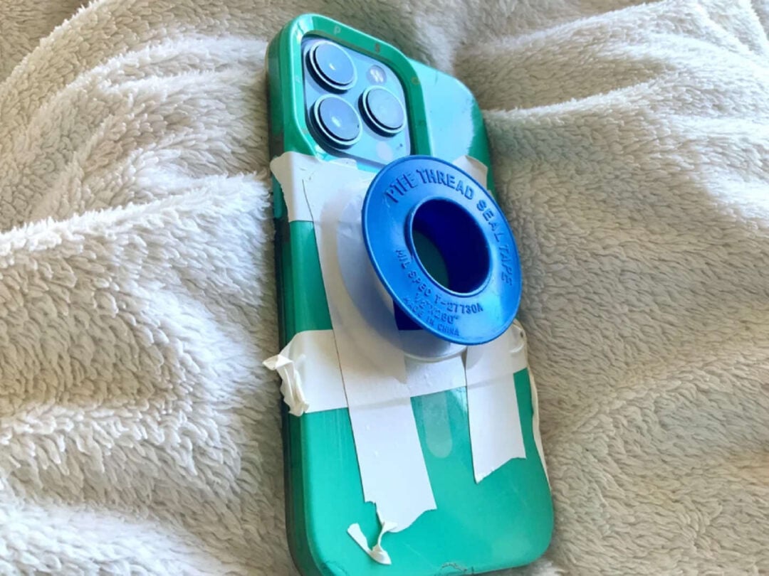 A green smartphone with a blue plastic spool taped to its back using white tape rests on a soft, light-colored blanket. The spool appears to be a roll of pipe thread seal tape.