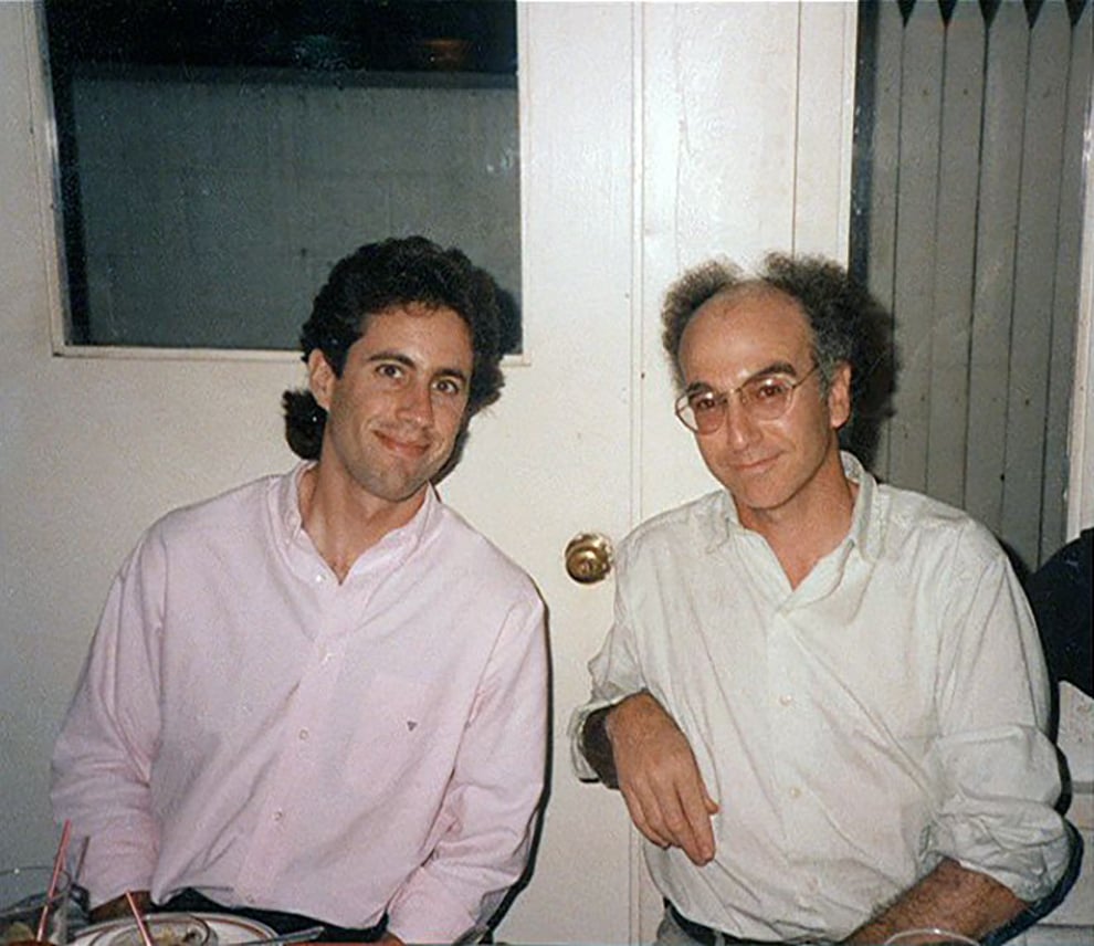 Two men in light-colored shirts are sitting next to each other at a table, smiling at the camera. There are drinks and plates in front of them and a closed white door behind them.