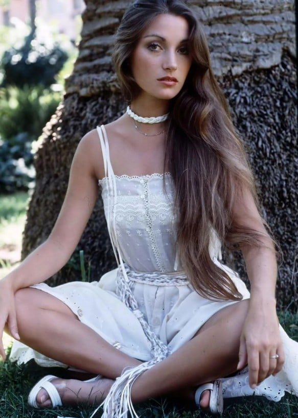 A young woman with long brown hair sits cross-legged outdoors in front of a tree, wearing a white sleeveless dress and sandals, with a choker necklace and a calm expression.