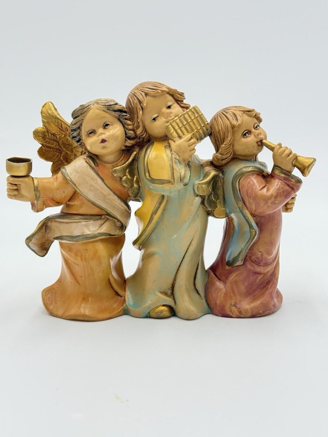 Three figurines of angels with wings stand side by side. One holds a cup, another plays panpipes, and the third plays a trumpet. They wear flowing robes in earthy tones and have serene facial expressions.