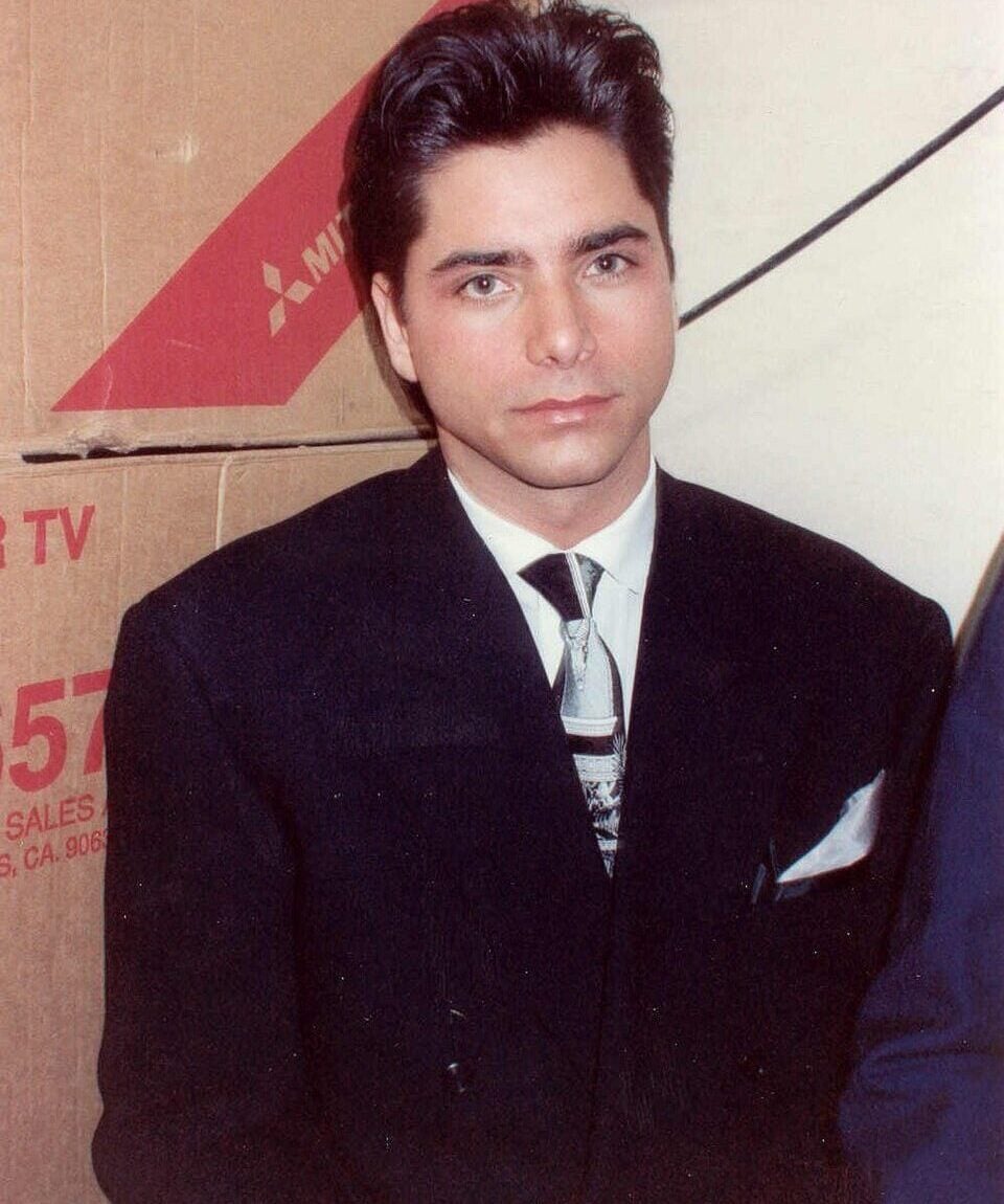 A young man with dark hair wearing a black suit, white shirt, and patterned tie stands in front of cardboard boxes and looks at the camera with a neutral expression.