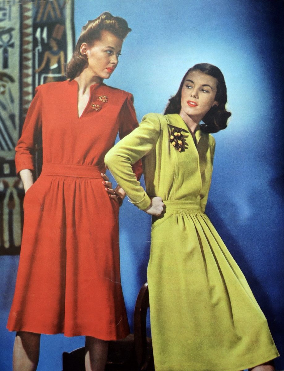 Two women pose against a blue background, wearing 1940s-style dresses—one in red, one in yellow-green. Both dresses have long sleeves, pleated skirts, and brooches pinned to the left side of the chest.