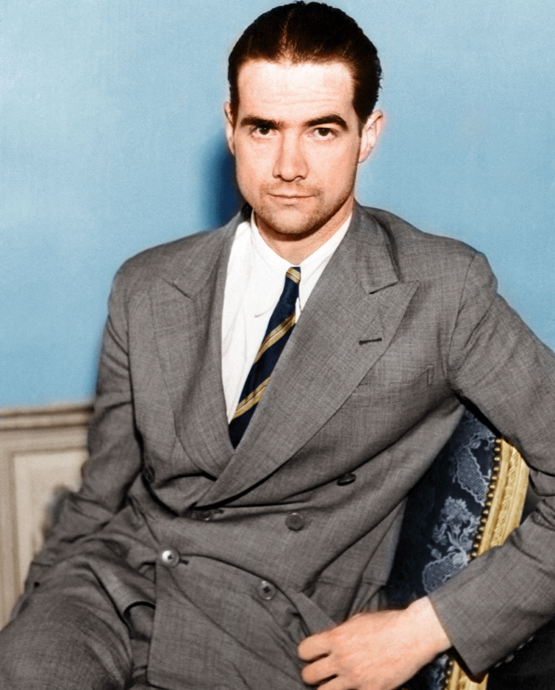 A young man with slicked-back dark hair wears a light gray suit, white shirt, and striped tie. He sits on a blue upholstered chair against a pale blue background, looking seriously at the camera.