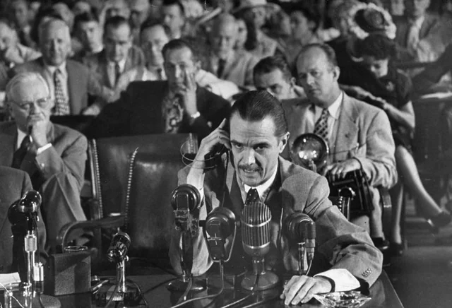 A man in a suit speaks into multiple microphones at a table during a formal hearing, with a large audience seated behind him, some appearing attentive and others taking notes.