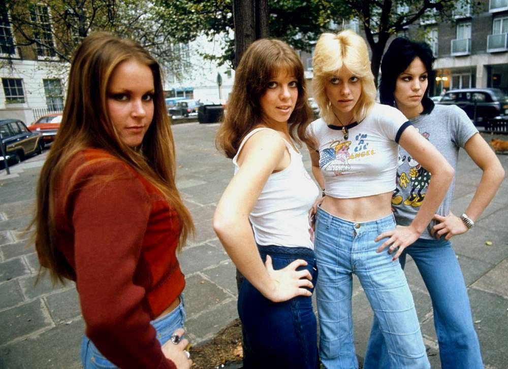 Four young women stand confidently outdoors on a sidewalk, posing with hands on hips. They wear 1970s-style clothing, including jeans and casual tops, with trees and parked cars visible in the background.