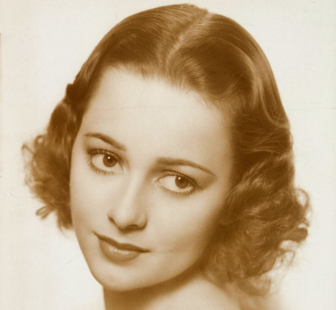 A sepia-toned portrait of a young woman with short, wavy hair and soft features, gazing slightly past the camera with a gentle expression. Her shoulders are bare, and the background is plain and light.