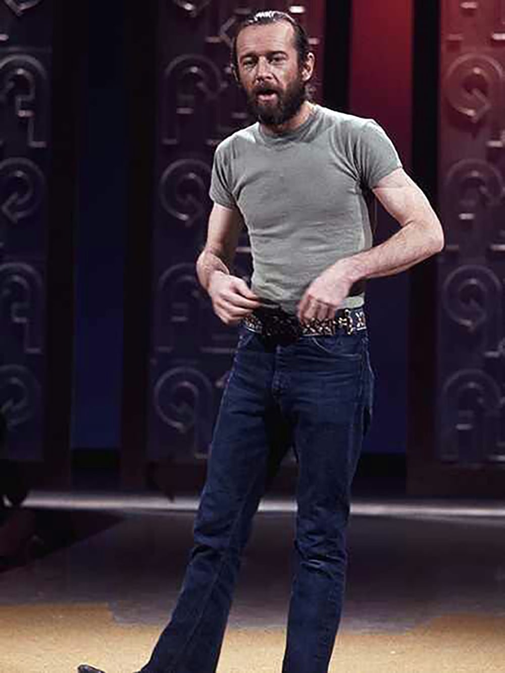 A man with a beard and mustache stands on stage wearing a fitted gray t-shirt, blue jeans, and a studded belt. The background features patterned panels and dramatic lighting.