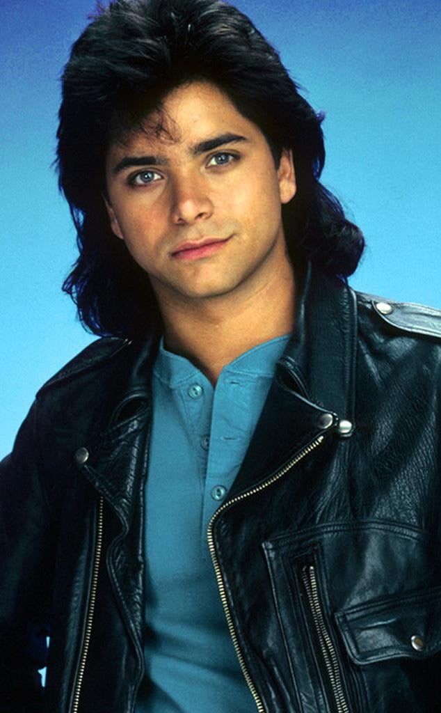 A young man with dark, wavy hair wears a black leather jacket over a blue shirt and looks confidently at the camera against a solid blue background.