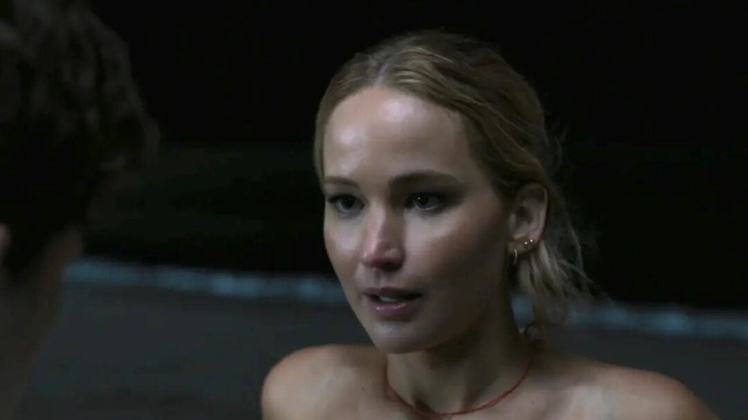 A woman with blonde hair tied back stands outdoors at night, looking slightly to the side with a neutral expression. The background is dark and blurry, possibly a beach or shoreline.