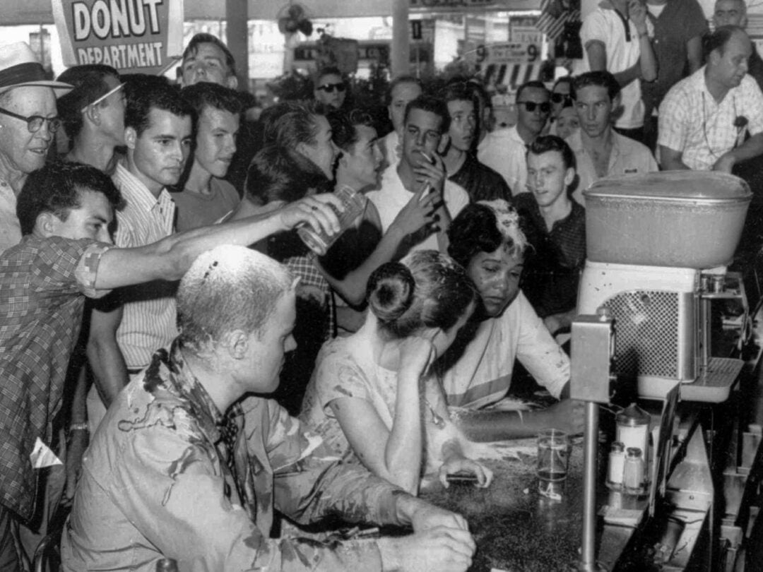 A group of white men harass and pour food on Black and white protesters sitting at a diner counter during a civil rights sit-in, with a "Donut Department" sign visible in the background.