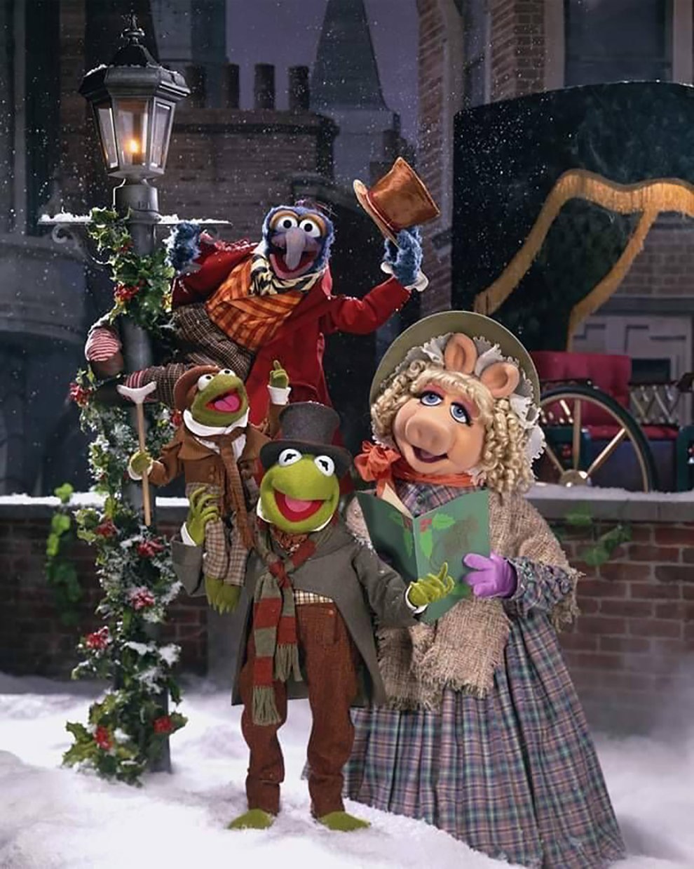 Four Muppets dressed in Victorian-era Christmas outfits stand in the snow by a decorated lamppost, caroling. There is snow on the ground and a horse-drawn carriage in the background.