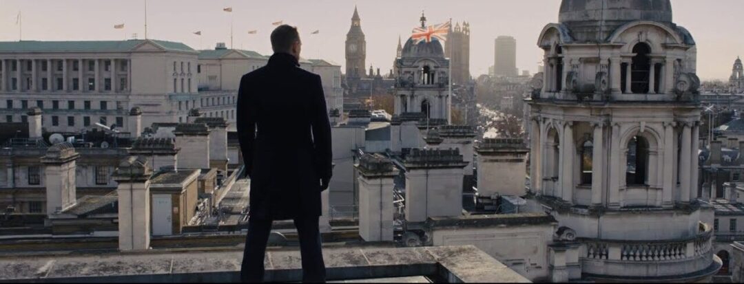 A person in a dark coat stands on a rooftop overlooking a cityscape with historic buildings, including Big Ben and the Union Jack flag, under a cloudy sky.